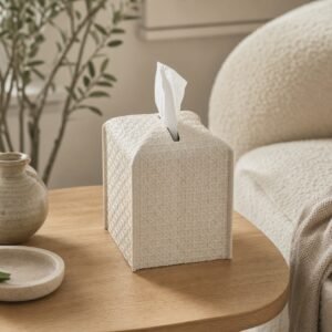 Beige rattan-style tissue box cover on a round wood table beside a cream chair